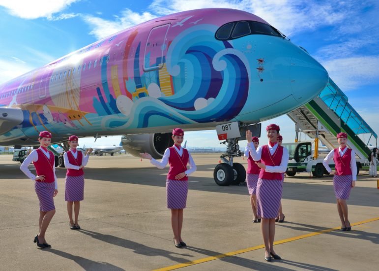 Wide-body airliner in a colourful wave-themed livery with cabin crew posing on the apron